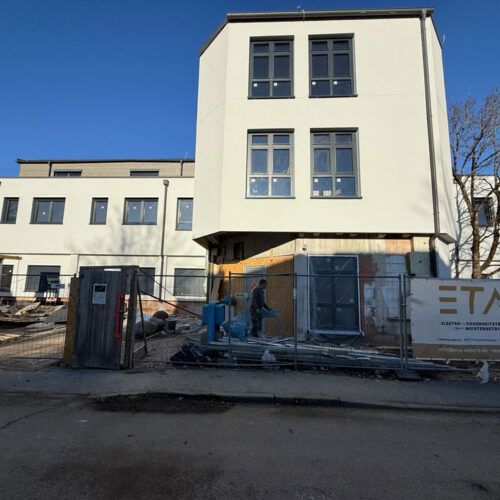 Eine Baustelle mit weißem Gebäude, mehreren Fenstern; klarer Himmel im Hintergrund.