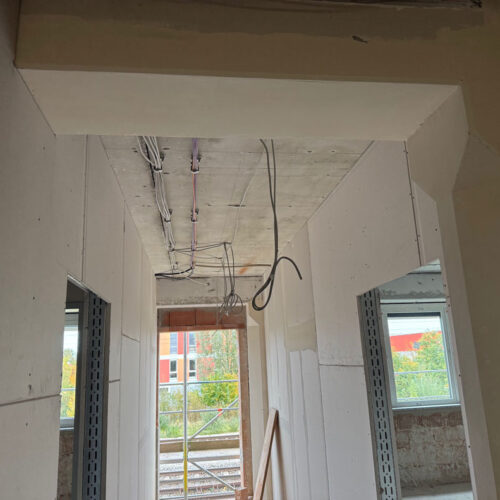 Rohbauflur mit sichtbaren Kabeln an der Decke, weiße Wände, Fenster mit Ausblick auf Baugerüst.