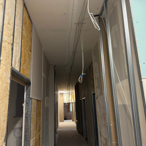 Baustelle mit nackten Wänden, gelbe Isolierung sichtbar, lose Kabel hängen von der Decke.