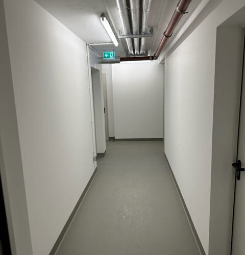 Ein leerer, langer Flur mit weiß gestrichenen Wänden und sichtbaren Lüftungsrohren an der Decke.