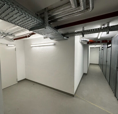 Ein leerer, heller Flur mit grauen Böden und Wänden; Metallrohre und -schränke sind sichtbar.