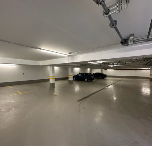 Eine leere Tiefgarage mit zwei geparkten Autos; Betonsäulen und weiß-graue Wände.