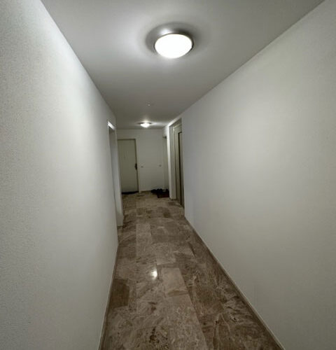 Langer, leerer Flur mit reflektierendem Marmorboden und zwei Deckenleuchten.