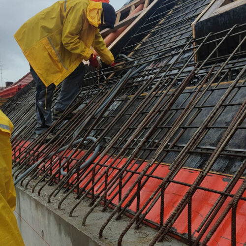 Arbeiter mit gelber Regenjacke auf Baustelle, konzentriert, Stahlgerüst, regenverhangener Himmel.