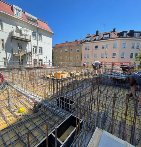 Bauarbeiter errichten Stahlgerüst auf Baustelle, helle Wohnhäuser im Hintergrund, klarer blauer Himmel.