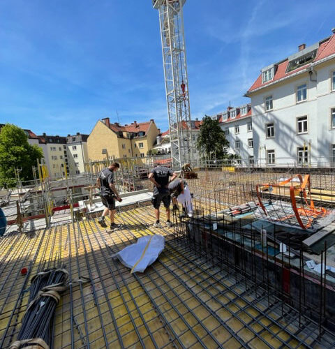 Drei Personen auf einer Baustelle, lachend, mit Bewehrungsstahl und Baukran im Hintergrund, sonnig.