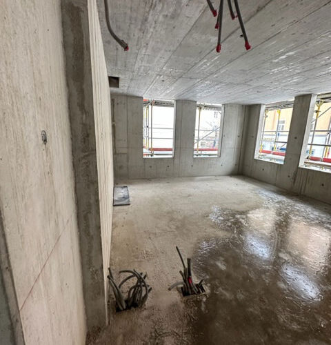 Rohbau eines Raumes mit Betonwänden und -boden, freiliegenden Kabeln und Fenstern im Hintergrund.