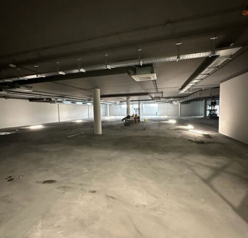 Ein leerer, beleuchteter Raum im Keller mit grauem Betonboden und Säulen.