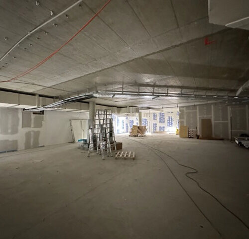 Leere Baustelle mit grauem Boden, Leiter, Kabeln und im Hintergrund Kartons und Baumaterial.