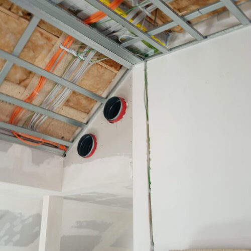 Decke mit sichtbaren Rohren und Kabeln, zwei rote Lüftungsöffnungen, unfertiger Raum, weiße Wände.