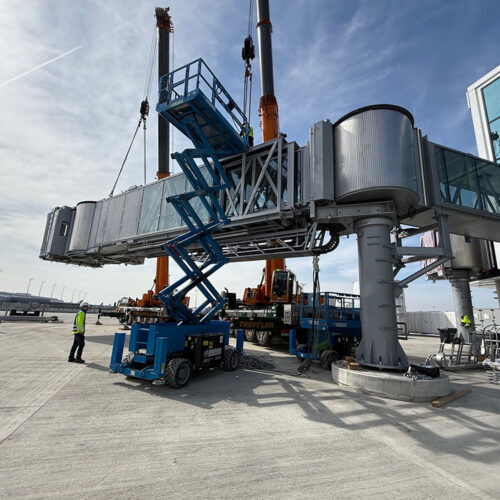 Bauarbeiter auf einem Flughafen installieren eine Fluggastbrücke mit Kränen und Hebebühne.