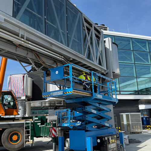 Arbeiter auf blauer Hebebühne, reparieren graue Gebäudestruktur vor Glasfassade, bei sonnigem Wetter.