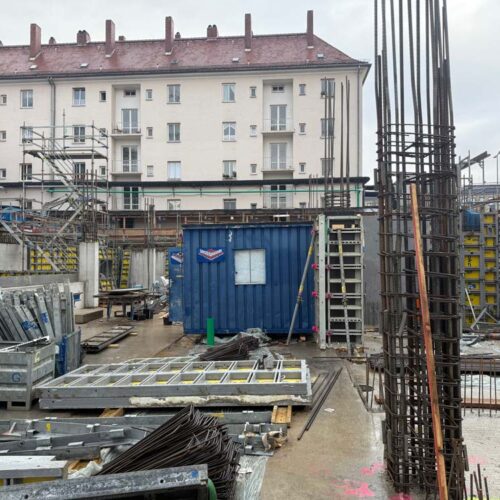 Baustelle mit Stahlträgern, Container, Gerüsten, grauem Himmel und einem mehrstöckigen Gebäude.