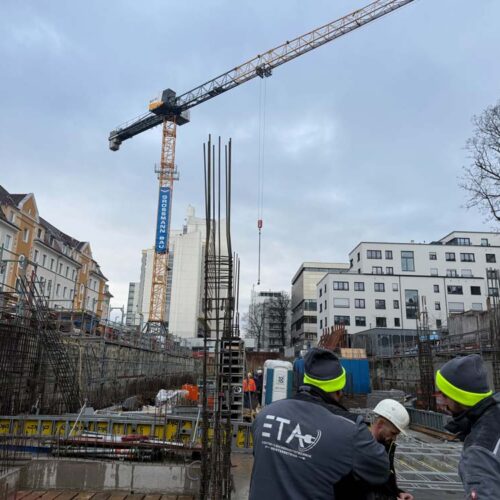 Bauarbeiter diskutieren auf Baustelle mit Kran und umgebenden Gebäuden, bedeckter Himmel.