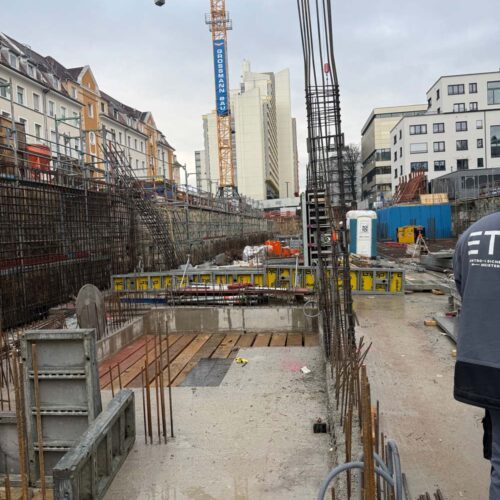 Baustelle mit Kran, Stahlgerüsten und Gebäuden im Hintergrund, bewölkter Himmel.