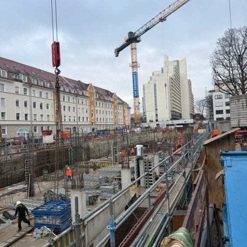 Baustelle mit Kränen und Arbeitern in Schutzkleidung; Gebäude und Bäume im Hintergrund, bewölkter Himmel.
