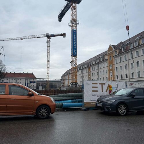 Baustelle mit Kränen und Autos auf nasser Straße. Umgebende Wohngebäude, bewölkter Himmel.