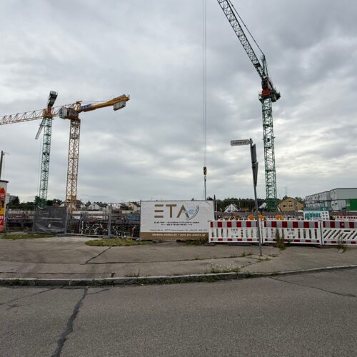 Baustelle mit zwei hohen Kränen, Bauzaun und bewölktem Himmel.