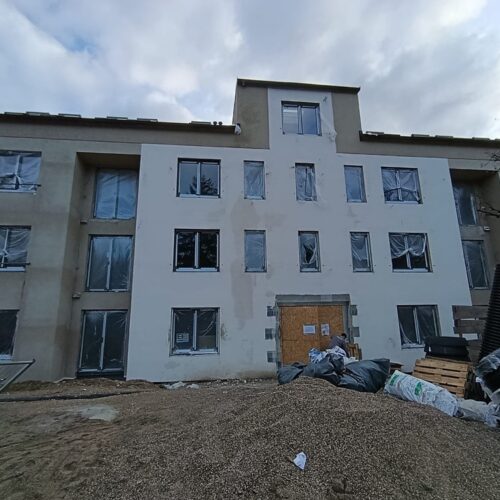 Ein  mehrstöckiges,  helles  Gebäude  im  Rohbau  mit  verschlossenen  Fenstern  und  Baumaterialien  davor