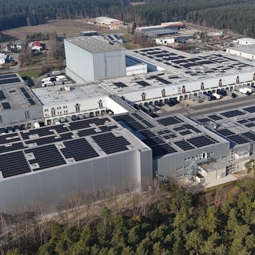 Luftaufnahme  einer  grauweissen  Fabrik  mit  schwarzen  Solarmodulen  im  Waldgebiet