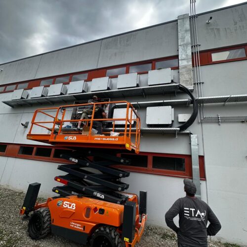 Zwei  Männer  wirken  konzentriert  an  grauer  Fassade  mit  orangefarbener  Hebebuehne